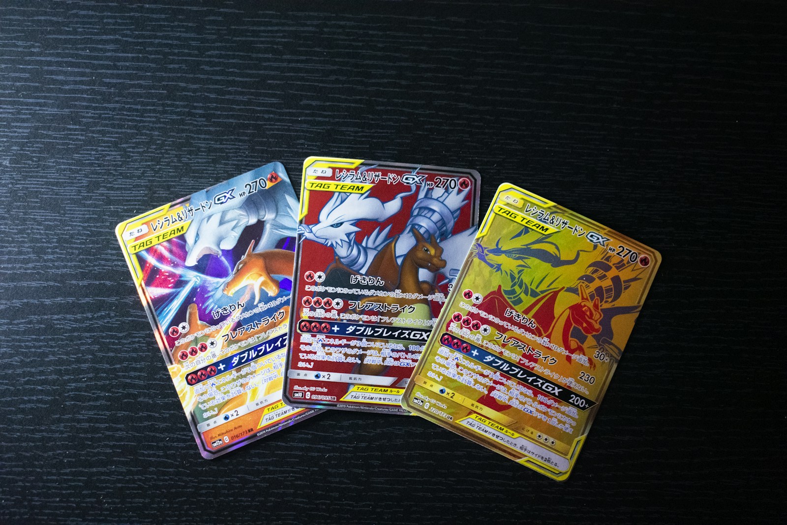 Tre carte giapponesi di Reshiram e Charizard GX con illustrazioni diverse su un tavolo scuro.